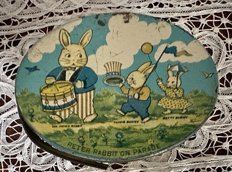 Puede incluir: Una fiambrera de metal vintage con una ilustraci&oacute;n colorida de Peter Rabbit y otros conejos en un desfile. Los conejos est&aacute;n vestidos con atuendos patri&oacute;ticos, uno tocando un tambor y otro sosteniendo un globo. El texto "Peter Rabbit on Parade" est&aacute; impreso en la parte inferior de la fiambrera.