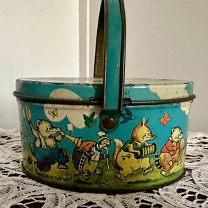 Puede incluir: Una fiambrera vintage de hojalata azul y blanca con asa. La fiambrera presenta una ilustraci&oacute;n caprichosa de animales tocando m&uacute;sica. El texto en la fiambrera dice "We Got To Pay The Rent".