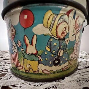 Puede incluir: Cubo de hojalata vintage con una ilustraci&oacute;n colorida de cuatro conejos. Los conejos llevan ropa y sostienen un globo rojo. El cubo tiene el texto "Mrs. Peter Rabbit's Garden" en la parte inferior.