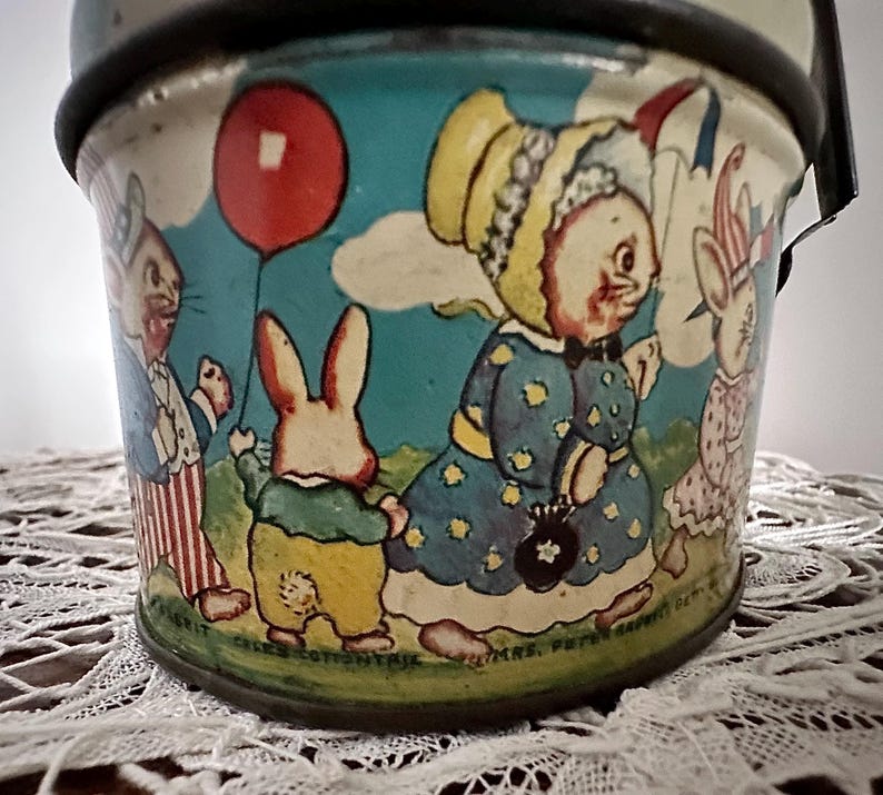 Puede incluir: Cubo de hojalata vintage con una ilustraci&oacute;n colorida de cuatro conejos. Los conejos llevan ropa y sostienen un globo rojo. El cubo tiene el texto "Mrs. Peter Rabbit's Garden" en la parte inferior.