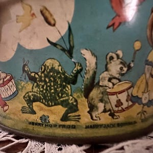 Puede incluir: Cubo de hojalata vintage con fondo azul e ilustraciones coloridas de una rana, una ardilla, un p&aacute;jaro y una ni&ntilde;a. El cubo tiene el texto "Henry Hop Frog" y "Happy Jack Squirrel" en la parte inferior.