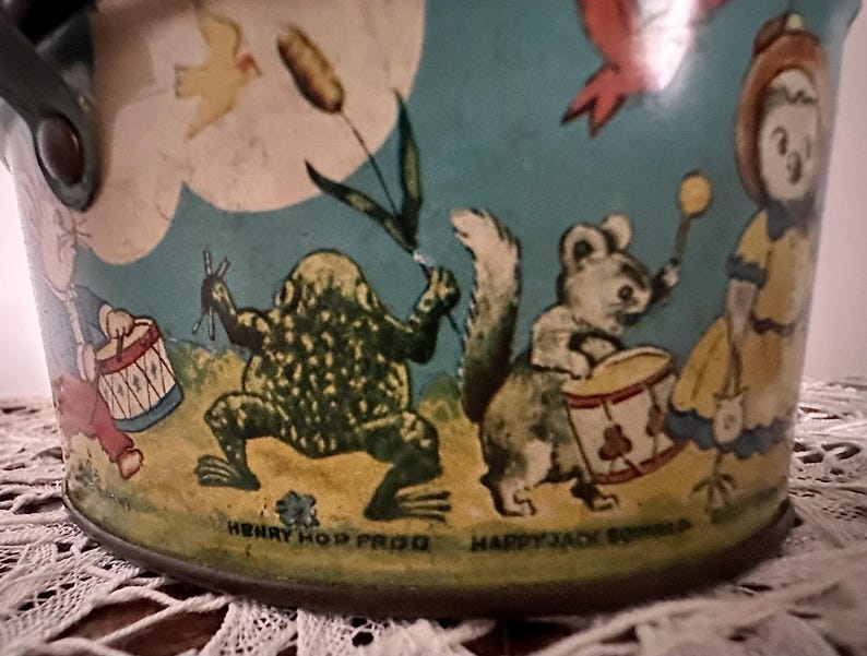 Puede incluir: Cubo de hojalata vintage con fondo azul e ilustraciones coloridas de una rana, una ardilla, un p&aacute;jaro y una ni&ntilde;a. El cubo tiene el texto "Henry Hop Frog" y "Happy Jack Squirrel" en la parte inferior.