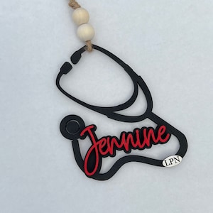 May include: Black stethoscope ornament with the name "Jennine" in red lettering and the abbreviation "LPN" in white lettering.