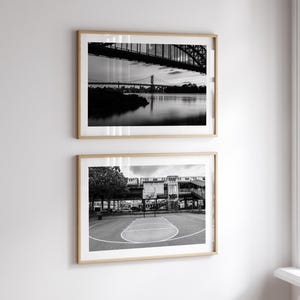 May include: Two framed black and white photographs hang on a white wall. The top image depicts a bridge over water, while the bottom shows a basketball court. The frames are light-colored wood.