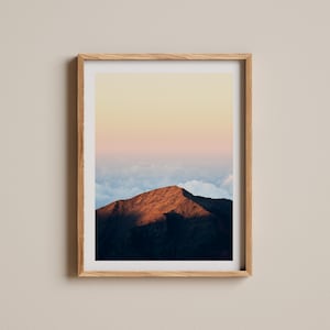 May include: A framed landscape print featuring a mountain peak bathed in warm sunlight. The mountain's reddish-brown surface contrasts with the dark shadows and the soft, pastel-colored sky above. The print is encased in a light-colored wooden frame.