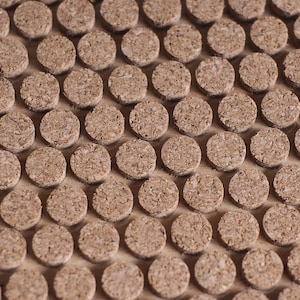 May include: A close-up view of a large quantity of round, brown cork circles arranged in a repeating pattern. The cork circles are all the same size and shape, and they are arranged in a staggered pattern.