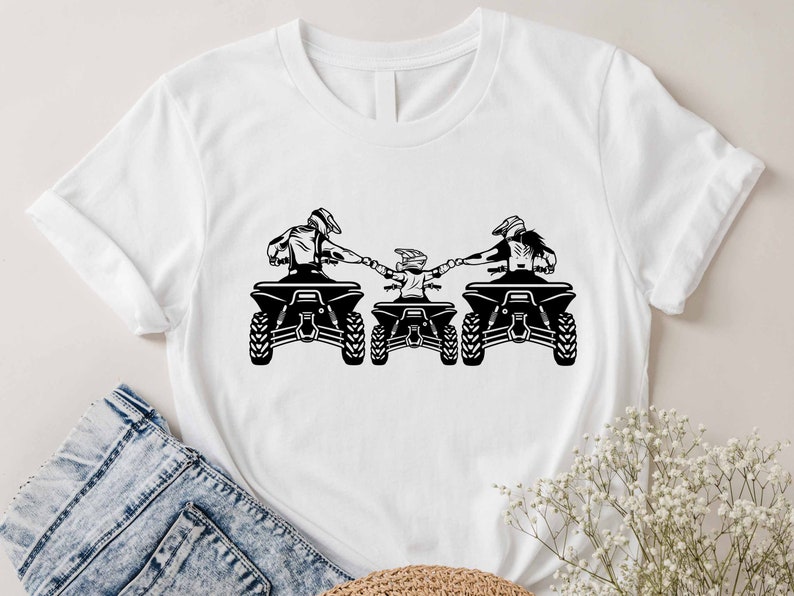 May include: A white t-shirt with a black silhouette of three people riding ATVs. The people are holding hands and the ATVs are facing forward.
