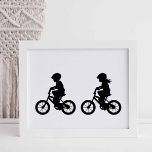 May include: Black and white silhouette print of two children riding bicycles. The print is framed in a white frame.