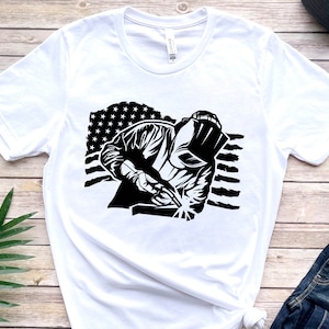 May include: White t-shirt with a black and white graphic of a welder working in front of an American flag.