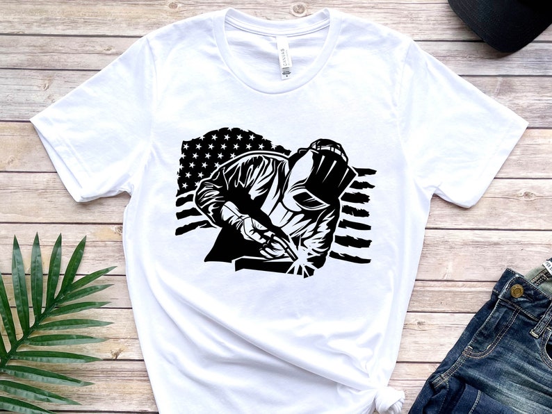 May include: White t-shirt with a black and white graphic of a welder working in front of an American flag.