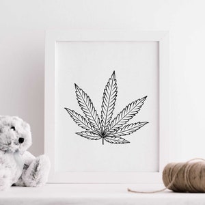 May include: A black and white line drawing of a marijuana leaf in a white frame. The frame is sitting on a white surface with a white teddy bear and a ball of twine.