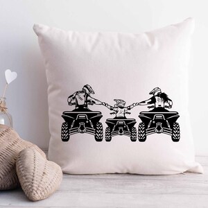 May include: A white throw pillow with a black silhouette of three people riding ATVs. The people are holding hands and are facing the same direction. The ATVs are all the same model and have a similar design.