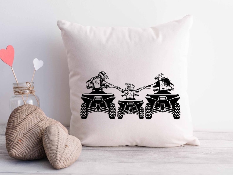 May include: A white throw pillow with a black silhouette of three people riding ATVs. The people are holding hands and are facing the same direction. The ATVs are all the same model and have a similar design.
