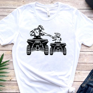 May include: White t-shirt featuring a black and white graphic of two people riding quad bikes. The riders are wearing helmets and holding onto the handlebars. The quad bikes are side-by-side and facing the same direction.