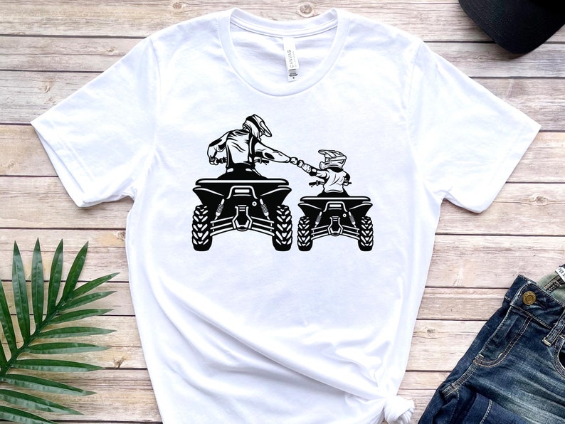 May include: White t-shirt featuring a black and white graphic of two people riding quad bikes. The riders are wearing helmets and holding onto the handlebars. The quad bikes are side-by-side and facing the same direction.