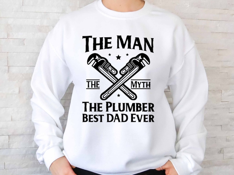 May include: White sweatshirt with black text that reads "The Man The Myth The Plumber Best Dad Ever". Two crossed wrench symbols are above the text.