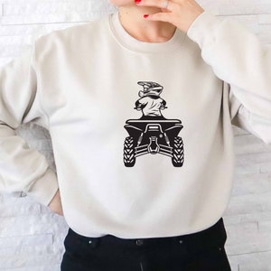 Puede incluir: Una sudadera blanca con un gr&aacute;fico negro de una persona conduciendo un ATV. La persona lleva un casco y gafas.