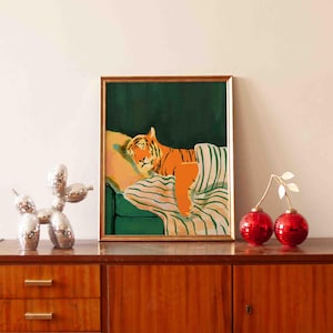 May include: A framed art print depicts an orange tiger resting on a green couch with a striped blanket. The artwork is in a gold frame and is displayed on a wooden cabinet. Decorative items include a disco dog sculpture and red ornaments.
