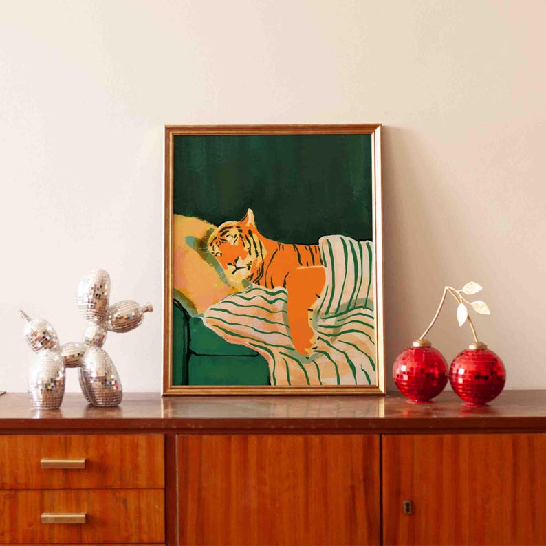May include: A framed art print depicts an orange tiger resting on a green couch with a striped blanket. The artwork is in a gold frame and is displayed on a wooden cabinet. Decorative items include a disco dog sculpture and red ornaments.