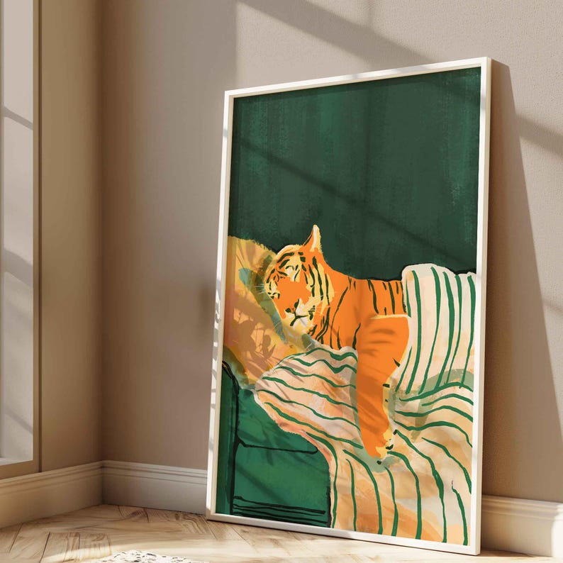 May include: A framed art print depicts an orange tiger resting on a green and striped sofa. The tiger is nestled on a yellow pillow and covered with a striped blanket. The background is a dark green color.