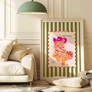 May include: A framed art print showing an orange and yellow tiger wearing a pink cowboy hat. The image is set within a pink postage stamp design, with the words "STAY WILD" above and "21&cent;" below. The background has green stripes.