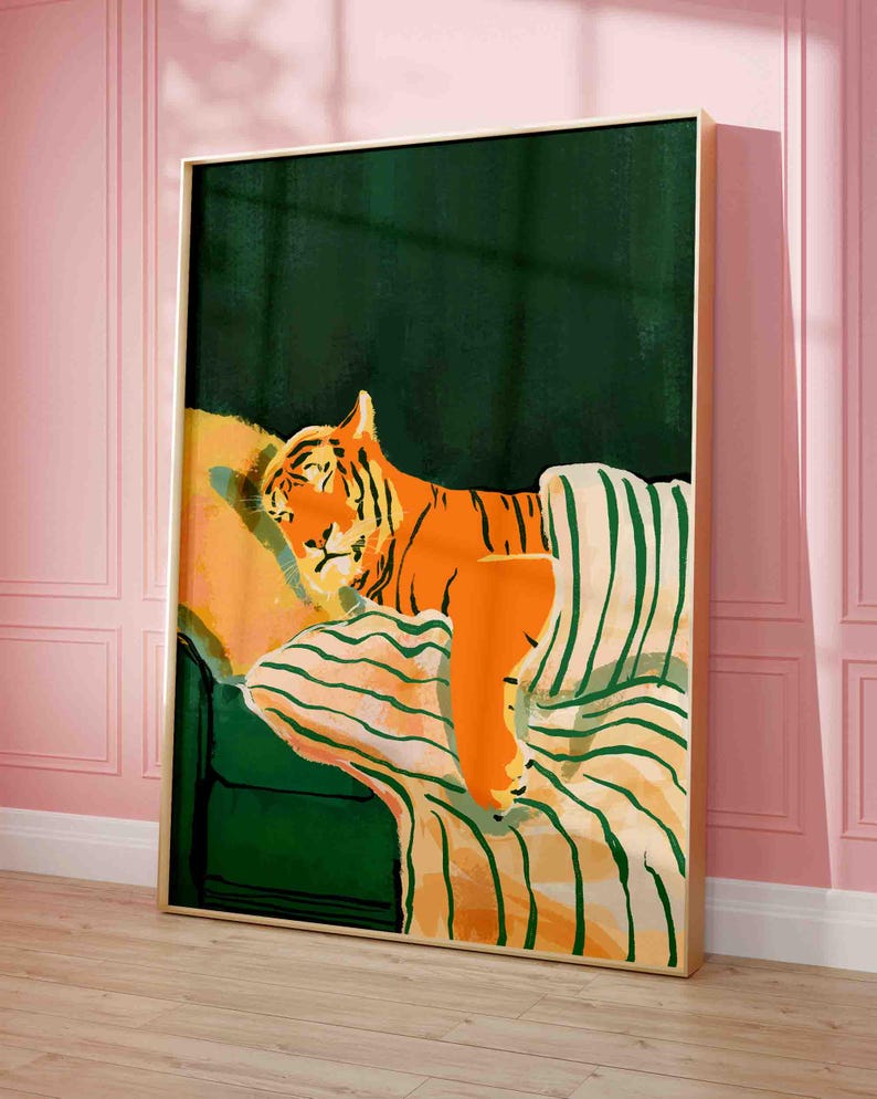 May include: A framed art print featuring an orange tiger resting on a green and striped sofa. The tiger has black stripes and is set against a dark green background. The artwork is in a gold frame and is displayed against a pink wall.