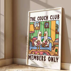 May include: A white framed print with a cartoon illustration of two ducks sitting on a couch. The couch is blue with a pink pillow. The ducks are holding a bucket of popcorn and a cup of coffee. The text on the print reads "The Couch Club Members Only".