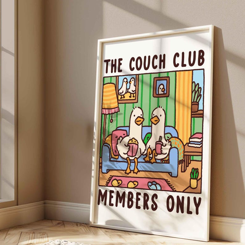 May include: A white framed print with a cartoon illustration of two ducks sitting on a couch. The couch is blue with a pink pillow. The ducks are holding a bucket of popcorn and a cup of coffee. The text on the print reads "The Couch Club Members Only".
