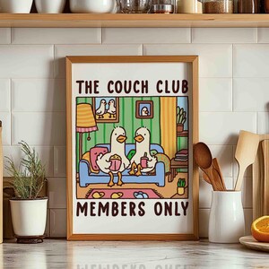 May include: A framed illustration of two cartoon ducks sitting on a couch with the text "The Couch Club Members Only". The ducks are wearing yellow and blue shirts and are holding snacks. The couch is blue with a yellow and orange patterned throw pillow.