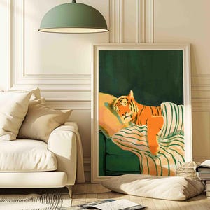 May include: A framed art print depicts a sleeping tiger on a green couch with striped cushions. The tiger is orange with black stripes, resting on a yellow pillow. The background is a dark green, and the frame is white.