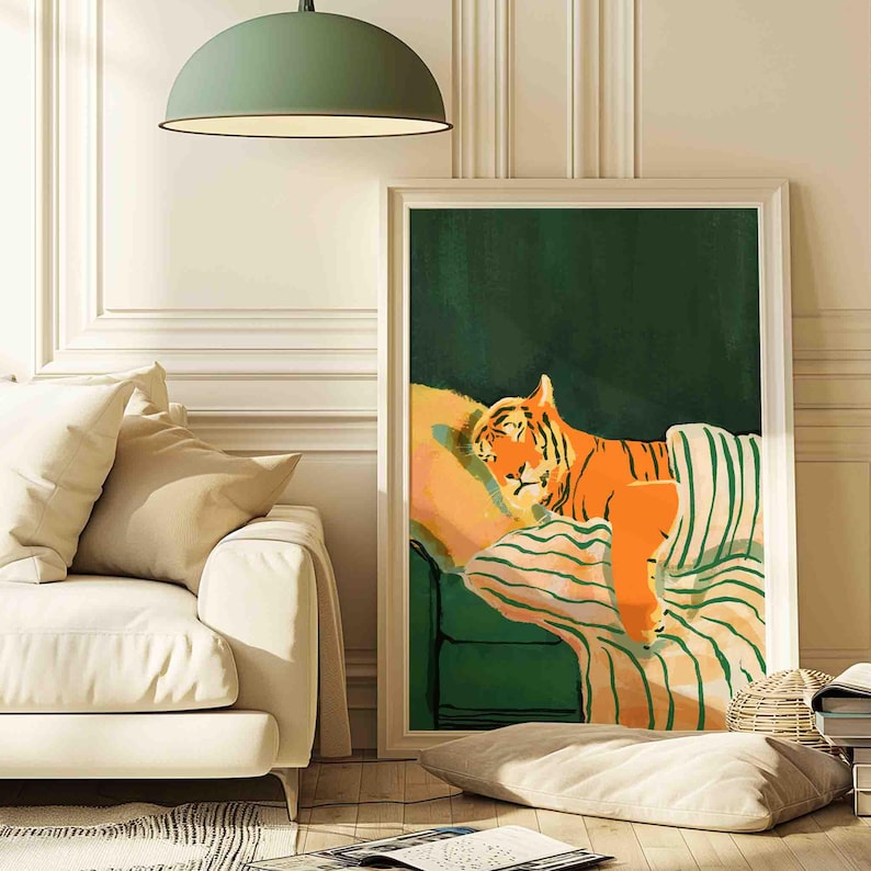 May include: A framed art print depicts a sleeping tiger on a green couch with striped cushions. The tiger is orange with black stripes, resting on a yellow pillow. The background is a dark green, and the frame is white.