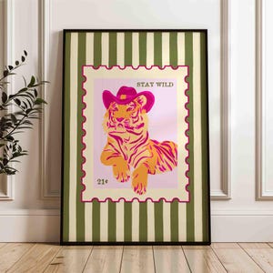 May include: Framed art print depicting a tiger in a pink cowboy hat, styled as a postage stamp. The tiger is orange and pink, with the words "STAY WILD" above. The frame has a green and cream striped pattern.