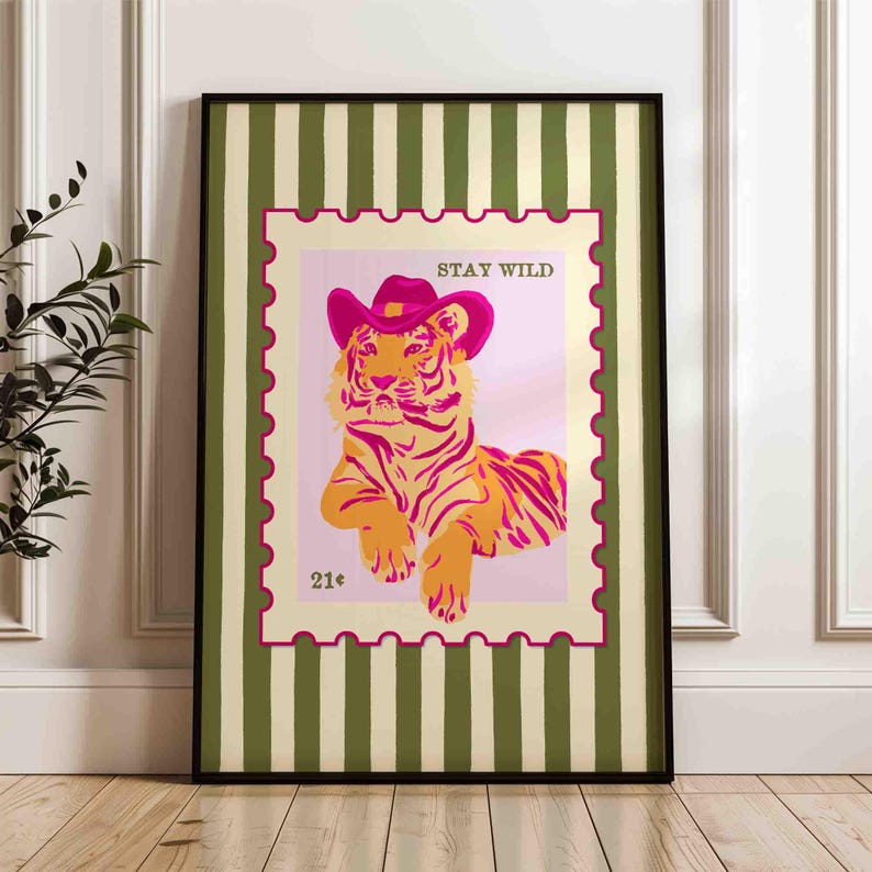 May include: Framed art print depicting a tiger in a pink cowboy hat, styled as a postage stamp. The tiger is orange and pink, with the words "STAY WILD" above. The frame has a green and cream striped pattern.