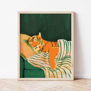 May include: A framed art print depicts an orange tiger resting on a green sofa with a striped blanket and pillow. The tiger is lying down, with its head resting on the pillow. The background is a dark green color.