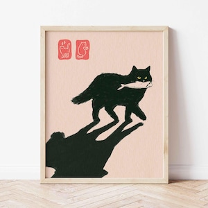 May include: A framed art print featuring a black cat carrying a fish in its mouth. The cat's shadow is cast below. Two red stamps are in the upper left corner. The background is a light pink color.