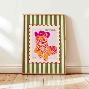 May include: Framed art print with a tiger wearing a pink cowboy hat, set on a pink background within a postage stamp design. The words "STAY WILD" are above the tiger. The frame has a green and white striped pattern.