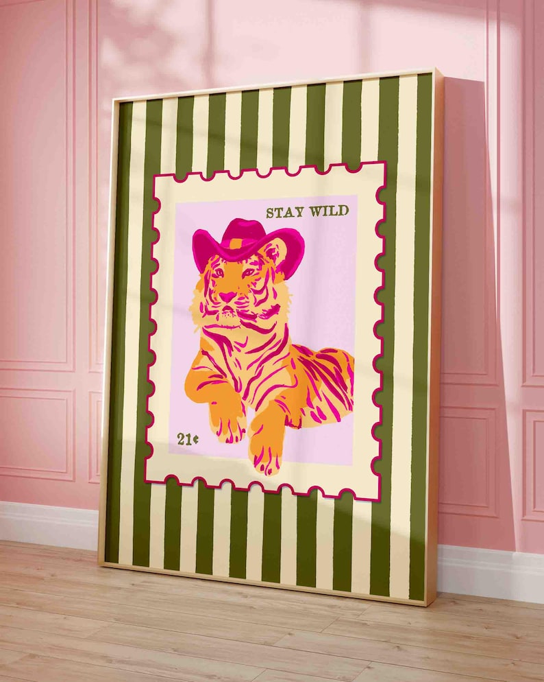 May include: A framed art print depicting a tiger wearing a pink cowboy hat. The tiger is orange and yellow with pink stripes, set against a pink background with the words "STAY WILD". The frame has a green and cream striped pattern.