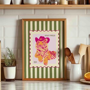 May include: A framed art print depicting a tiger in a pink cowboy hat, set within a postage stamp design. The words "STAY WILD" are at the top, with "21&cent;" at the bottom. The frame has a green and white striped background. The print is a decorative item.