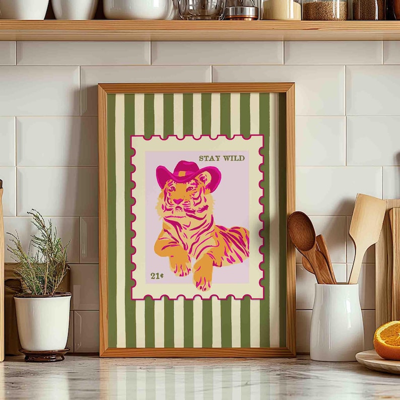 May include: A framed art print depicting a tiger in a pink cowboy hat, set within a postage stamp design. The words "STAY WILD" are at the top, with "21&cent;" at the bottom. The frame has a green and white striped background. The print is a decorative item.