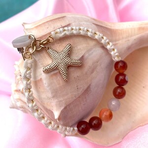 May include: A white cord bracelet with a gold starfish charm and red agate beads. The bracelet is displayed on a seashell.