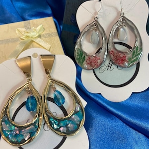 May include: Two pairs of dangle earrings. The first pair is gold with a blue teardrop crystal and iridescent shell accents. The second pair is silver with a white teardrop crystal and dried flowers.