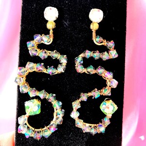 May include: A pair of statement earrings with a unique design. The earrings feature a gold-colored wire frame in a stylized shape, adorned with iridescent, faceted beads. Each earring has a clear, round stud at the top.