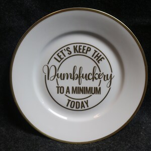 May include: White ceramic plate with a gold rim and a gold inscription that reads "Let's keep the Dumbfuckery to a minimum today."