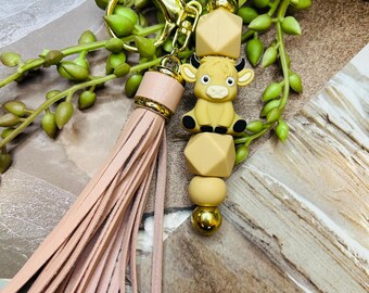 May include: A pink tassel keychain with a gold key ring and a brown cow charm. The charm is made of silicone and has a 3D design. The keychain is strung with brown and gold beads.