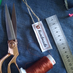 May include: A close-up of a sewing kit with a pair of rose gold scissors, a green awl, a spool of orange thread, a ruler, and a white tag with the word "handmade" stitched on it.