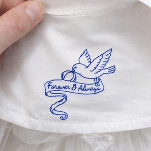 May include: White fabric with a blue embroidered dove carrying a ring and a banner that reads "Forever & Always." The fabric appears to be part of a wedding dress or similar garment.