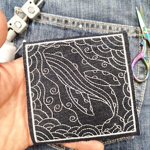 Könnte beinhalten: Quadratischer schwarzer Aufnäher mit einem weiß gestickten Wal-Design. Der Aufnäher hat einen weißen Rand und dekorative Wellen- und Wirbelmuster. Der Aufnäher wird in einer Hand gehalten, mit Jeans im Hintergrund.