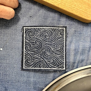 Könnte beinhalten: Quadratischer marineblauer Aufnäher mit einem weißen Wirbelwellenmuster, umrahmt von einem weißen Rand. Der Aufnäher liegt auf einer Denim-Oberfläche, was auf eine mögliche Verwendung für Kleidung oder Accessoires hindeutet.