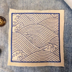 Può includere: Una toppa in tessuto quadrata con ricamo blu a linee che raffigura onde marine stilizzate. Il disegno è racchiuso all'interno di un bordo ricamato blu su uno sfondo beige chiaro, mostrando un motivo ripetitivo di onde che si infrangono.