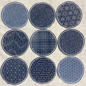 May include: Nine round denim coasters with white embroidered patterns. Each coaster features a unique design, including swirls, geometric shapes, and floral motifs. The coasters have a white stitched border and are arranged on a neutral fabric surface.